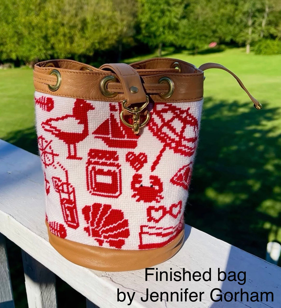 A bucket-style handbag with a red and white cross-stitch style nautical pattern, tan leather trim, and brass-tone hardware, photographed outdoors on a white railing with a green lawn in the background. Text at the bottom reads 'Finished bag by Jennifer Gorham.